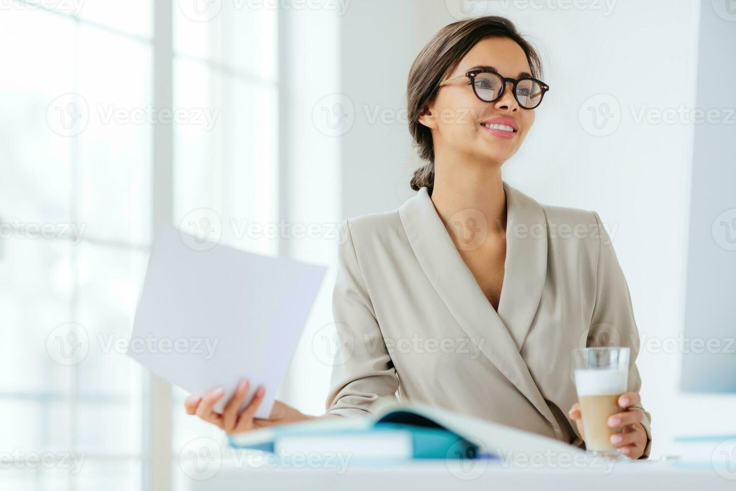 Cheerful businesswoman reads documents, smiles, drinks milkshake, makes project plan. Female executive manager in spectacles at coworking space photo