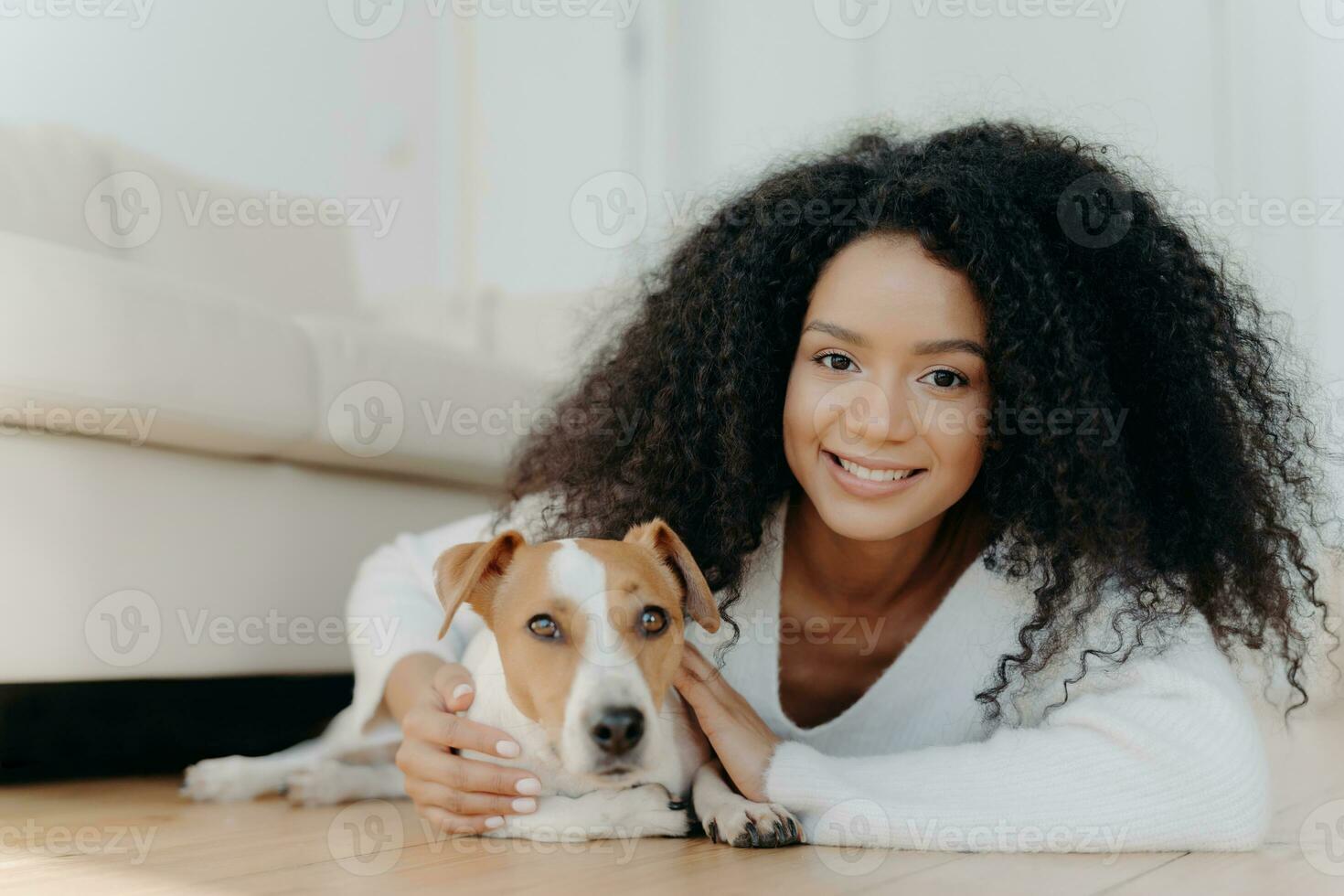 Pretty girl with Afro hair lies on living room floor, sharing pleasant emotions with newly adopted dog. Beloved pet in new apartment, creating beautiful moments together. photo