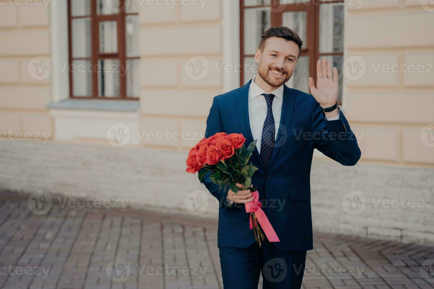 Happy man in blue suit waves and smiles, greets beloved woman with bouquet, standing alone outdoors. photo