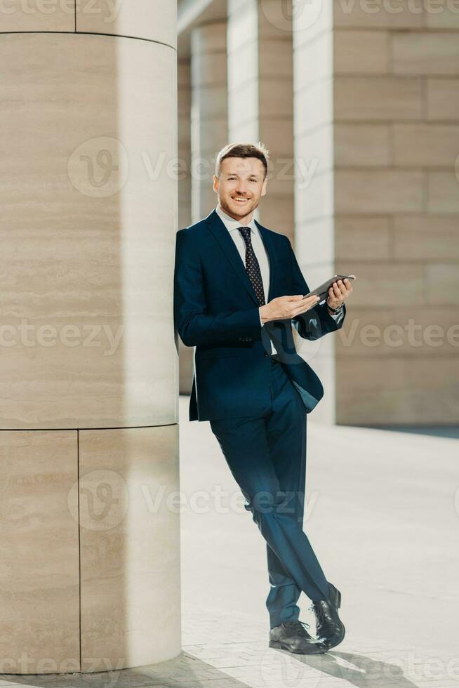 Photo of cheerful male dressed in expensive black suit, stands crossed legs, works on touch pad, stands in modern office interior, smiles positively at camera. Young male manager. Full length portrait
