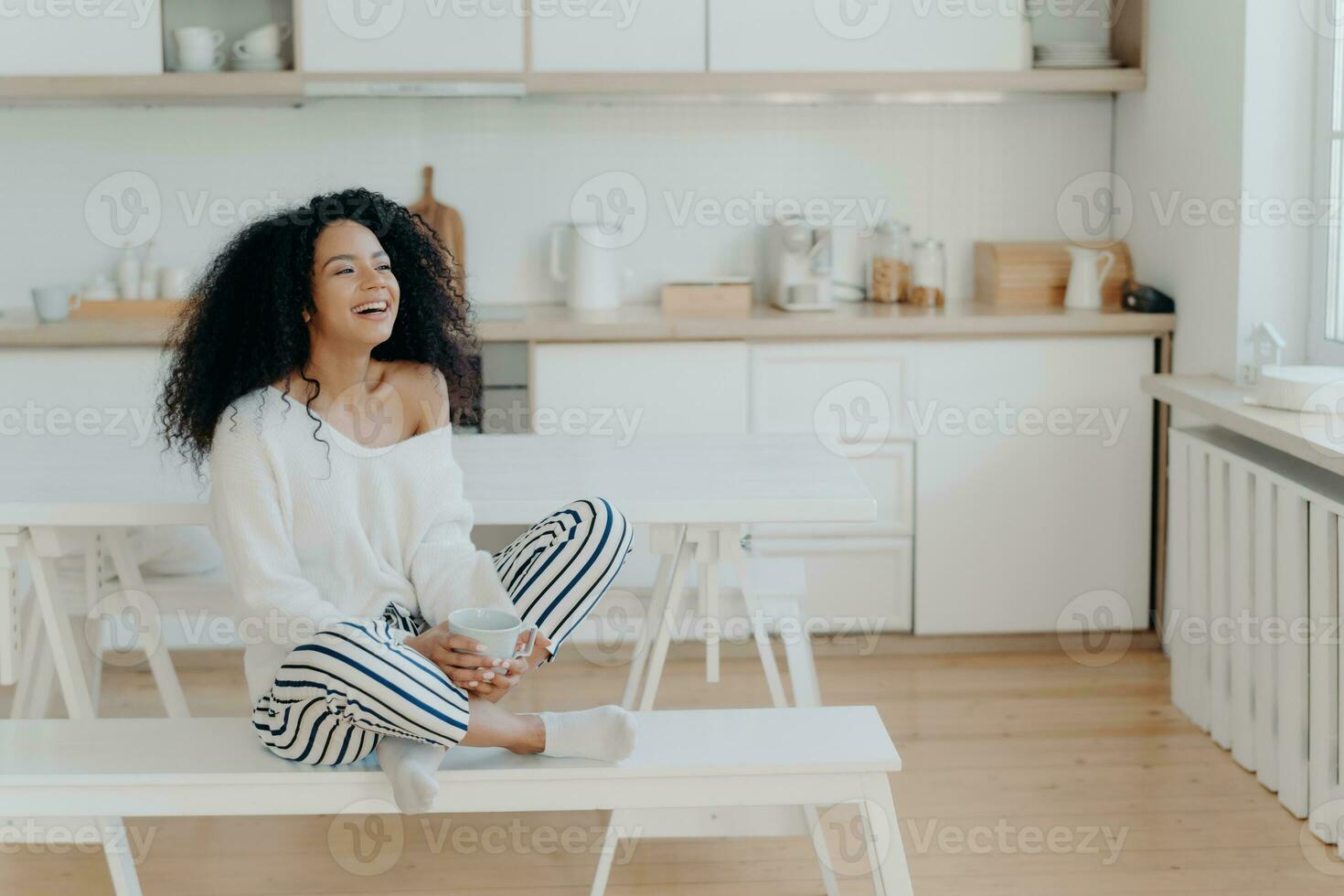 Overjoyed dark-skinned woman laughs, sips coffee, gazes out kitchen window in fashionable attire. Smiling lady enjoying hot beverage, home relaxation. photo