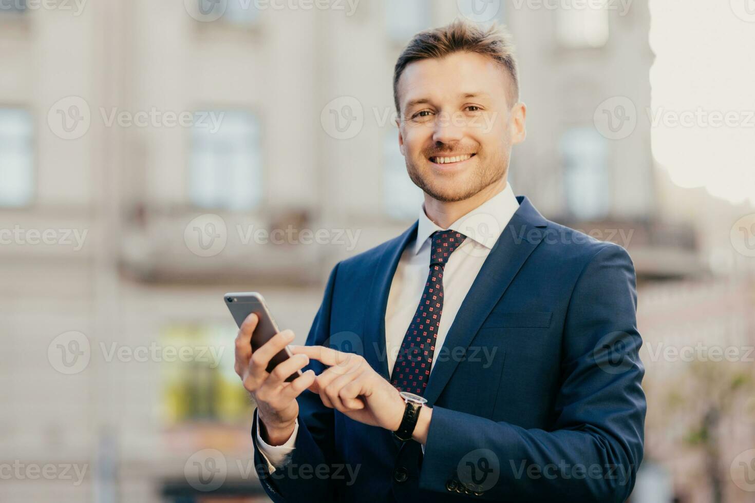 Satisfied businessman holds smart phone, reads email from investor, happy to recieve good news about business company, stands in urban territory, wears black suit and elegant tie. Technology concept photo