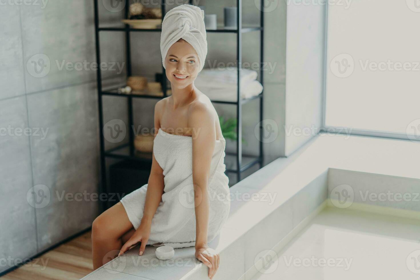 Relaxed woman with healthy skin smiles, towel-wrapped, uses cream, enjoys beauty routine, takes shower. Beauty, grooming concept. photo