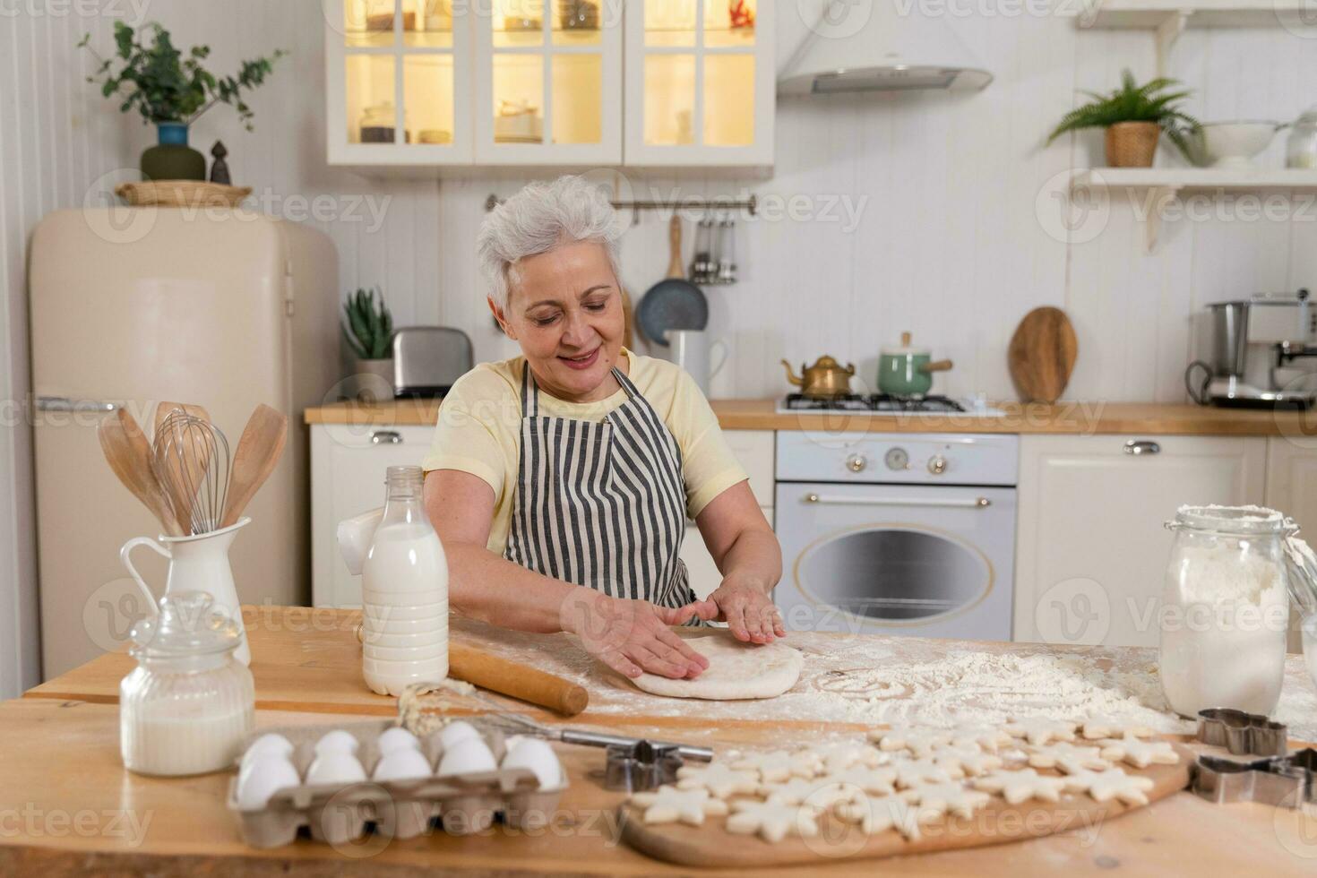 Happy senior woman cooking in kitchen. Stylish older mature gray haired lady grandmother knead dough bake cookies. Old grandma cook homemade food. Household housewife housework concept. photo