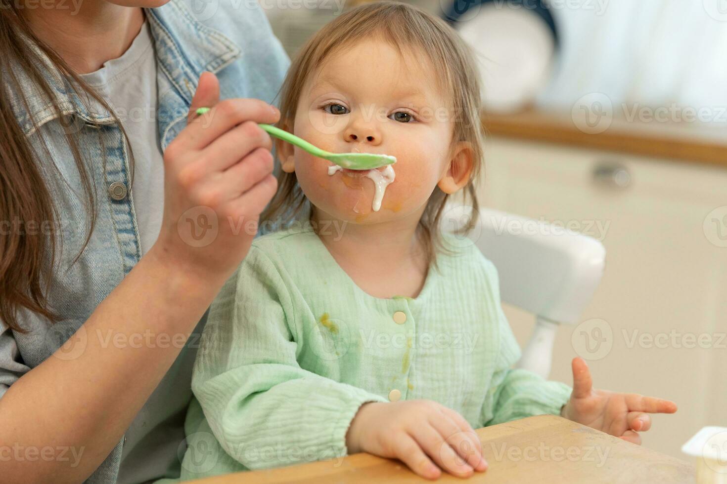 Happy family at home. Mother feeding her baby girl from spoon in kitchen. Little toddler child with messy funny face eats healthy food at home. Young woman mom giving food to kid daughter. photo