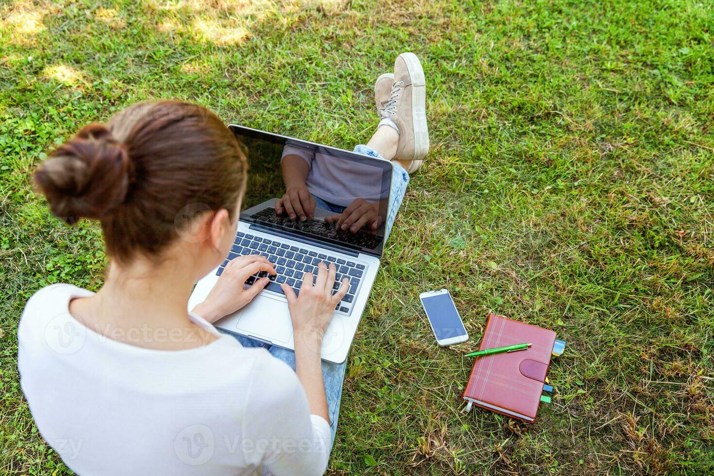 Young woman sitting on green grass lawn in city park working on laptop pc computer. Freelance business concept photo