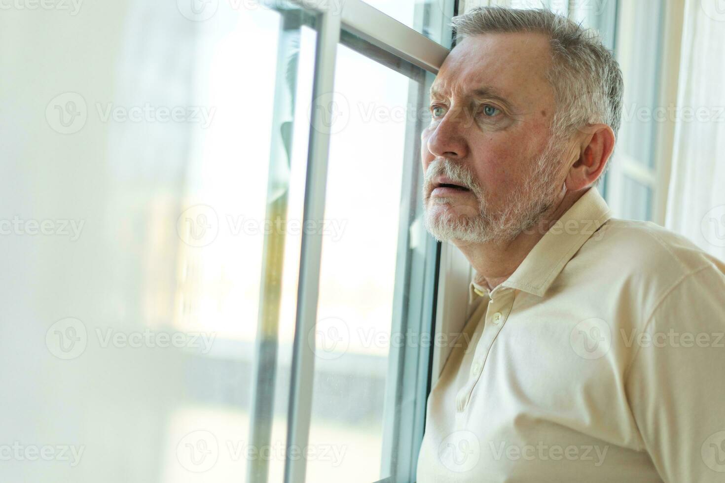 Sad tired lonely disappointed older senior man at home alone. Unhappy grandfather looking sadly outside window experiencing grief relative death bad news. Stressed elder man suffering from loneliness. photo