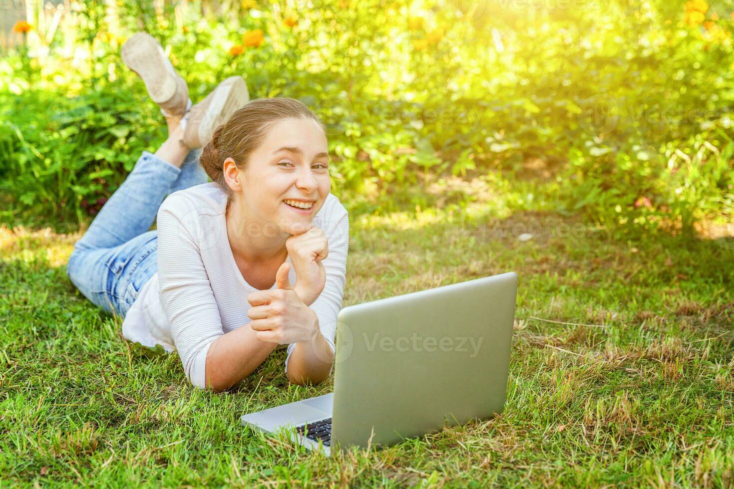 Young woman lying on green grass lawn in city park working on laptop pc computer. Freelance business concept photo