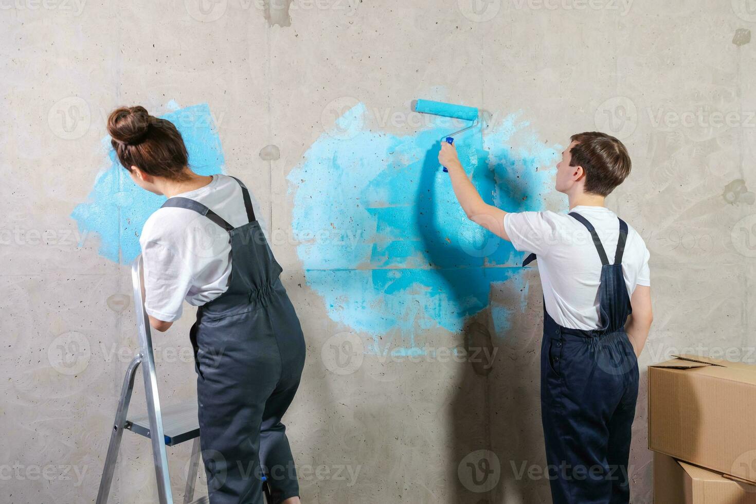 House decoration DIY renovation concept. Happy couple in empty room of new home painting wall together. Husband and wife holding paint roller painting wall with blue color paint in new house. photo