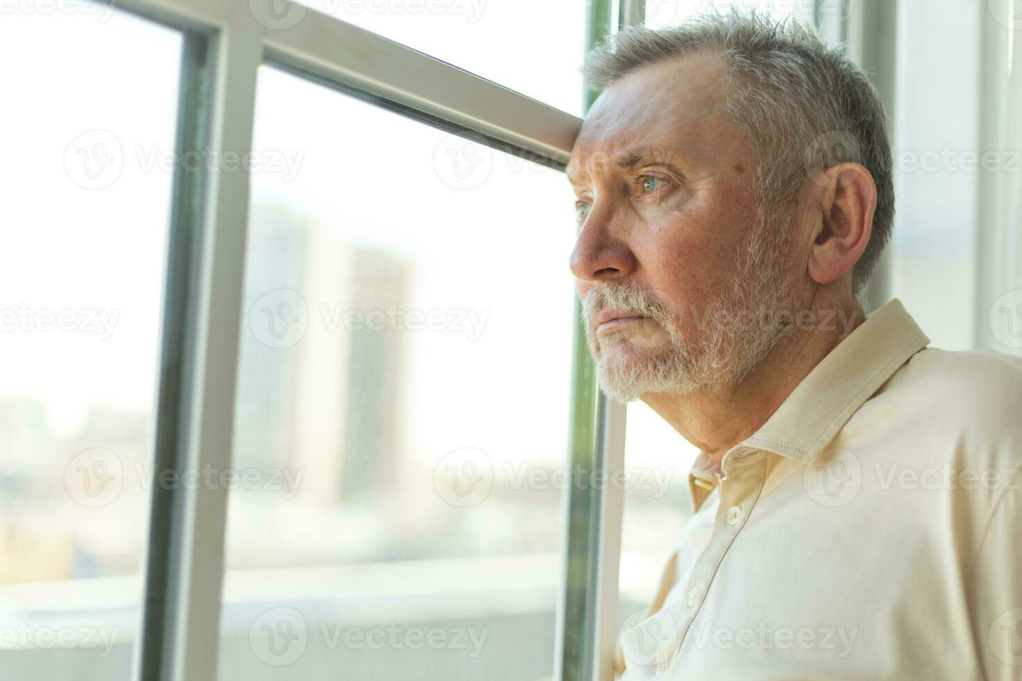 Sad tired lonely disappointed older senior man at home alone. Unhappy grandfather looking sadly outside window experiencing grief relative death bad news. Stressed elder man suffering from loneliness. photo
