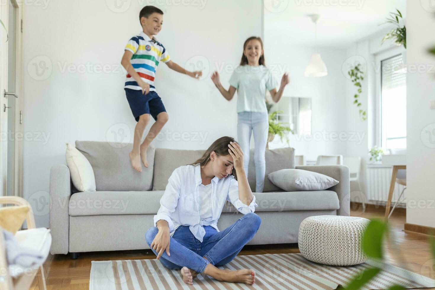 Tired young mother sitting on the floor and working with laptop and documents while little kids and jumping on the sofa and having fun and making noise photo