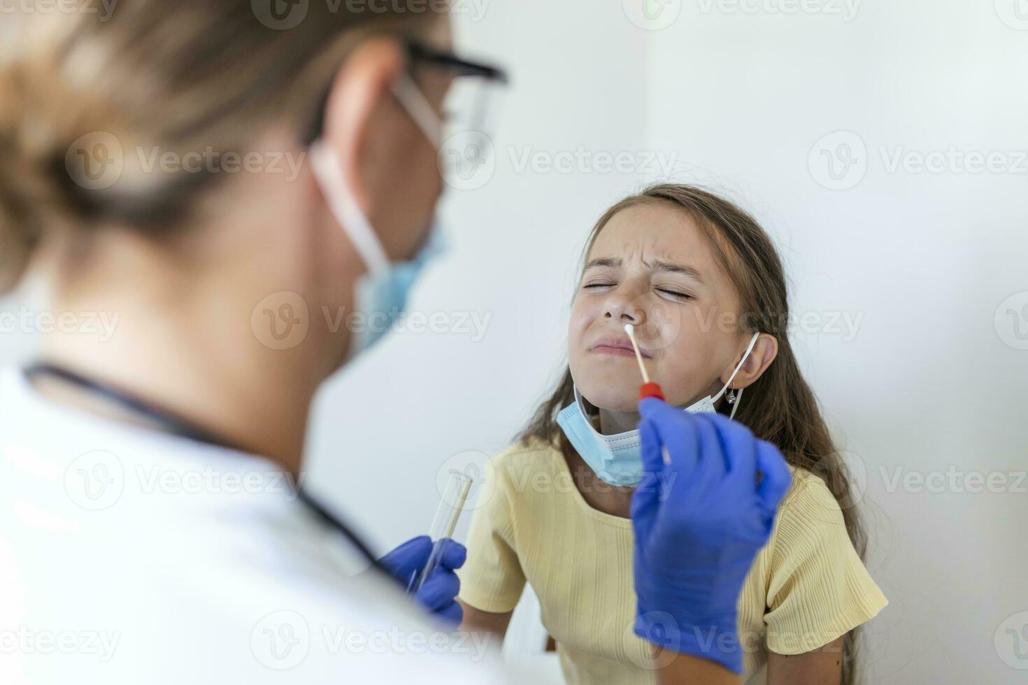 Nurse performing a nose swab test on a little child. Girl going through PCR testing due to COVID-19 pandemic. Female doctor using cotton swab while PCR testing small girl photo