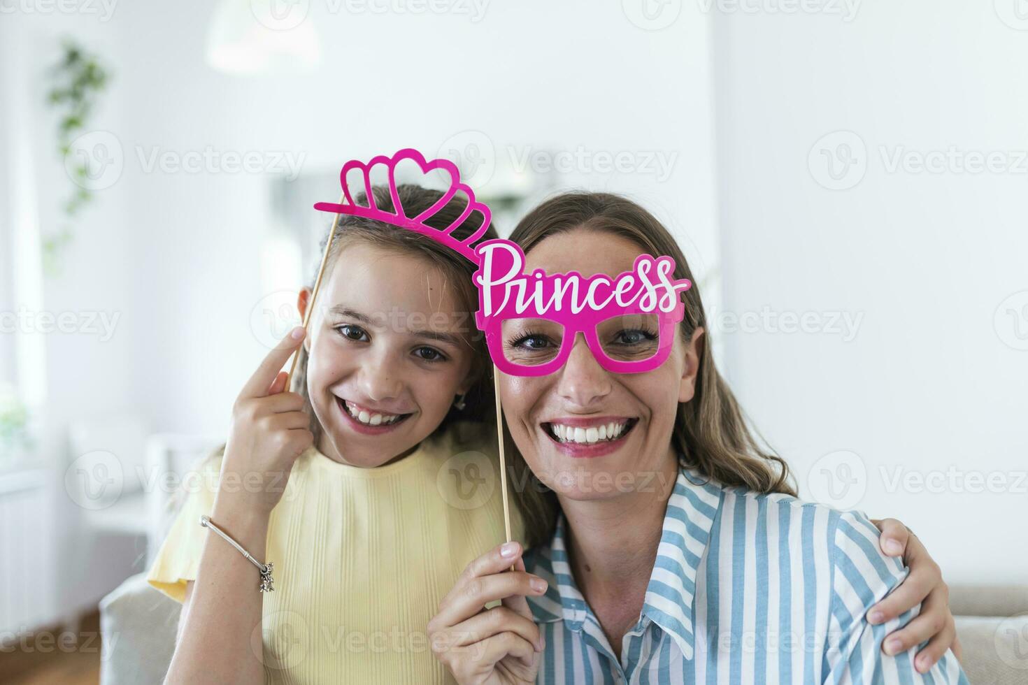 Funny family on a background of bright wall. Mother and her daughter girl with a paper accessories. Mom and child are holding paper crown on stick. photo