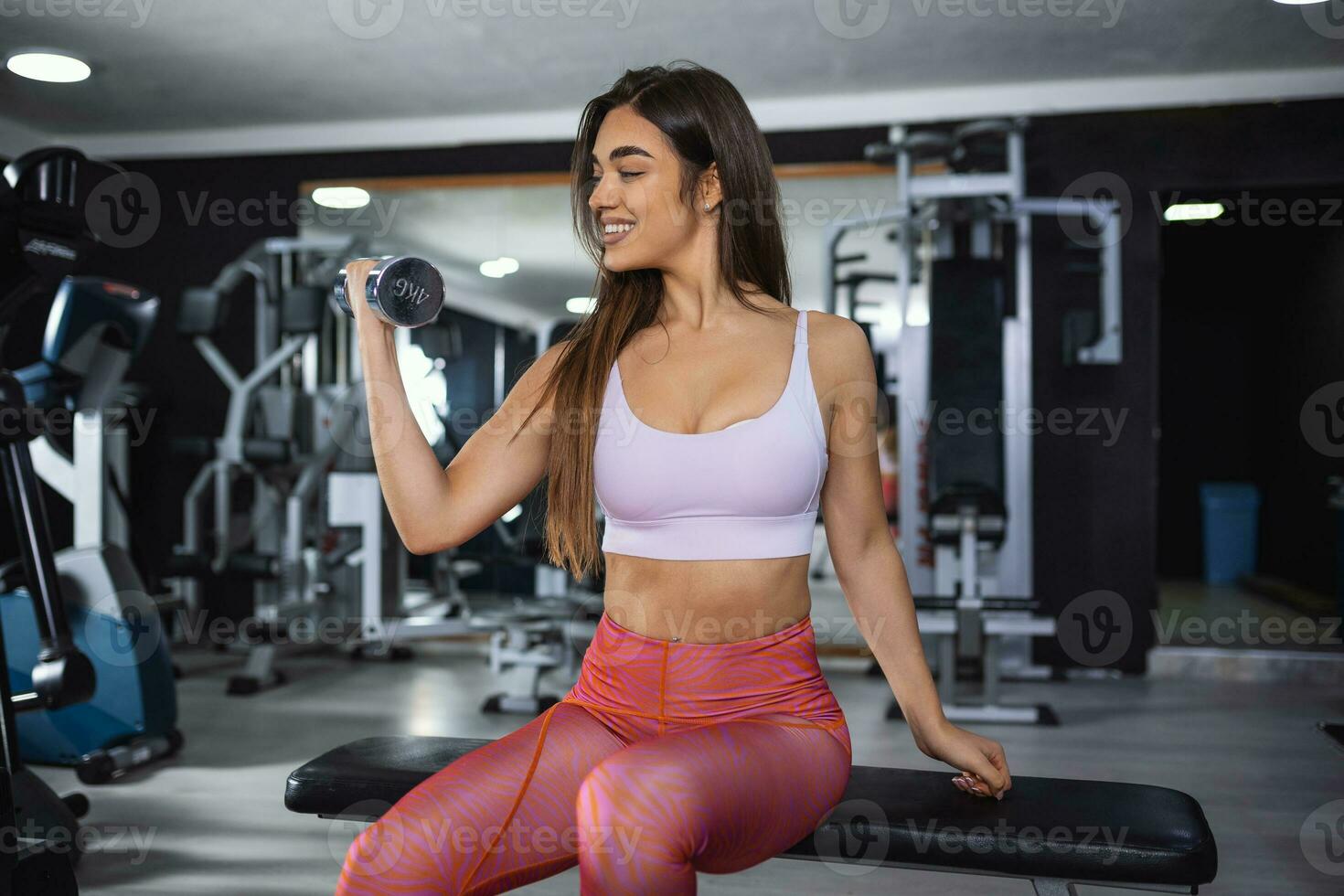 Beautiful athletic muscular woman pumps up the muscles by one arm lifts dumbbell exercise on bench in fitness gym. Young sport girl gains strong physical muscle well by weight lifted in fitness studio photo