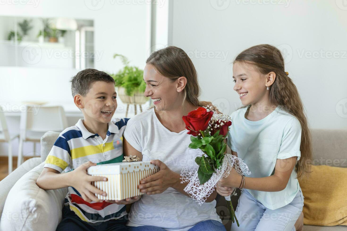 Happy children giving gift an flowers to mother. Happy Mothers Day. Children boy and girl congratulate smiling mother, give her flowers bouquet of roses and a gift box during holiday celebration photo