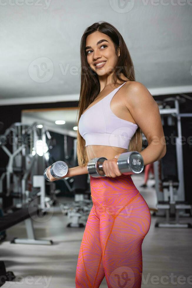 ajuste fuerte joven mujer levantamiento pesos trabajando fuera con pesas en pie con su lado a el cámara flexionando su brazo en el gimnasia. foto