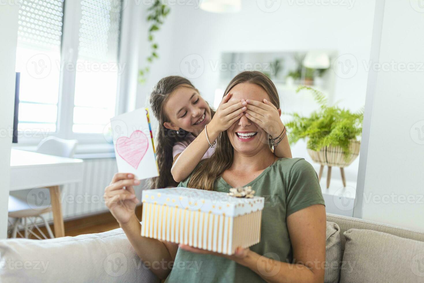 Cheerful little girl with gift box and card surprising and congratulating happy mom on mother day at home. Happy Mothers Day photo