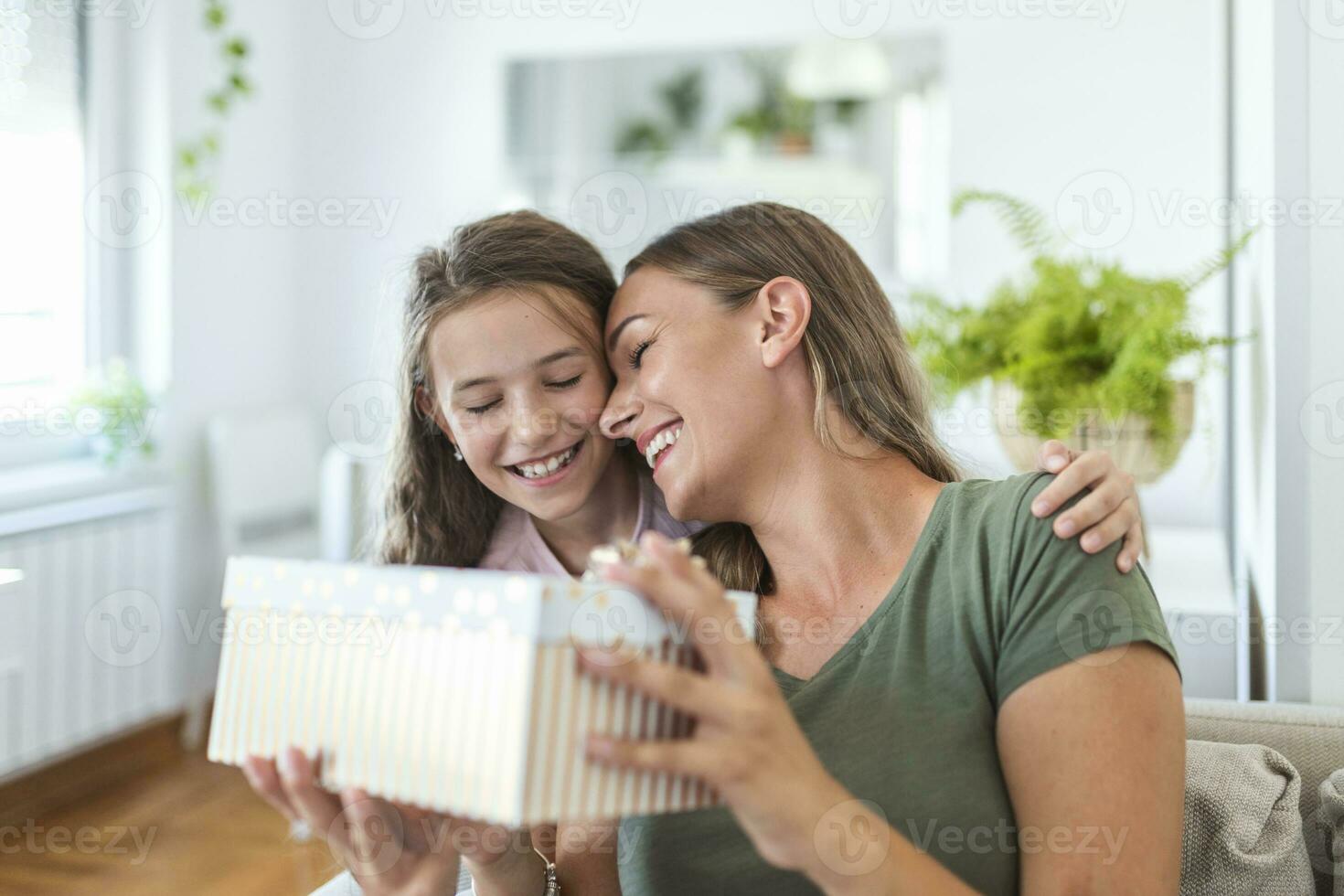 Young woman and girl at home celebrating mother's day sitting on sofa daughter hugging mother kissing cheek mom laughing joyful holding gift box photo