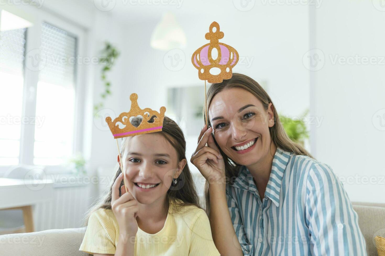 Funny family on a background of bright wall. Mother and her daughter girl with a paper accessories. Mom and child are holding paper crown on stick. photo