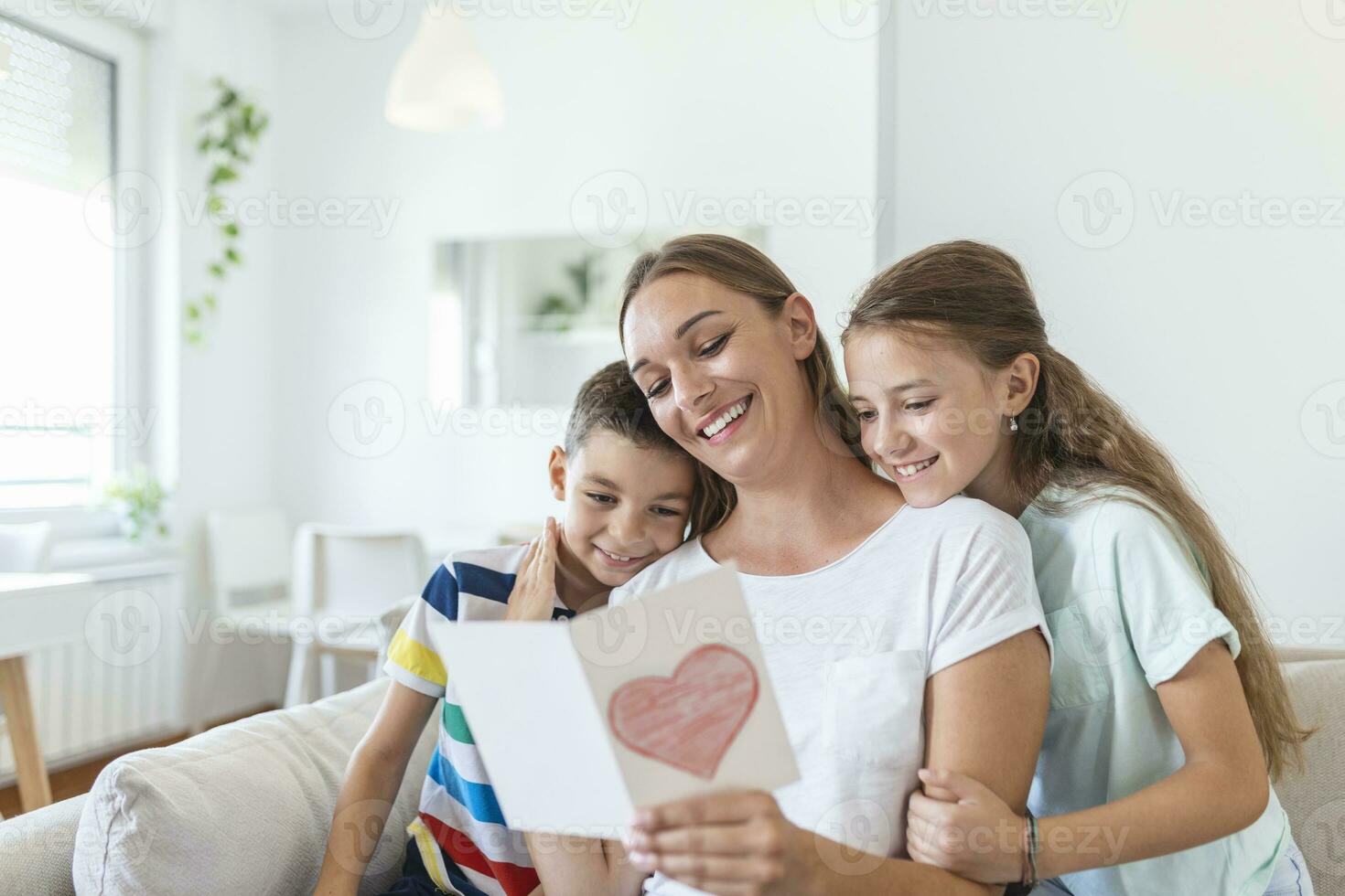 Joyful young mother and little children sitting on sofa and embracing while reading wishes and congratulations with holiday in presented postcard at home photo