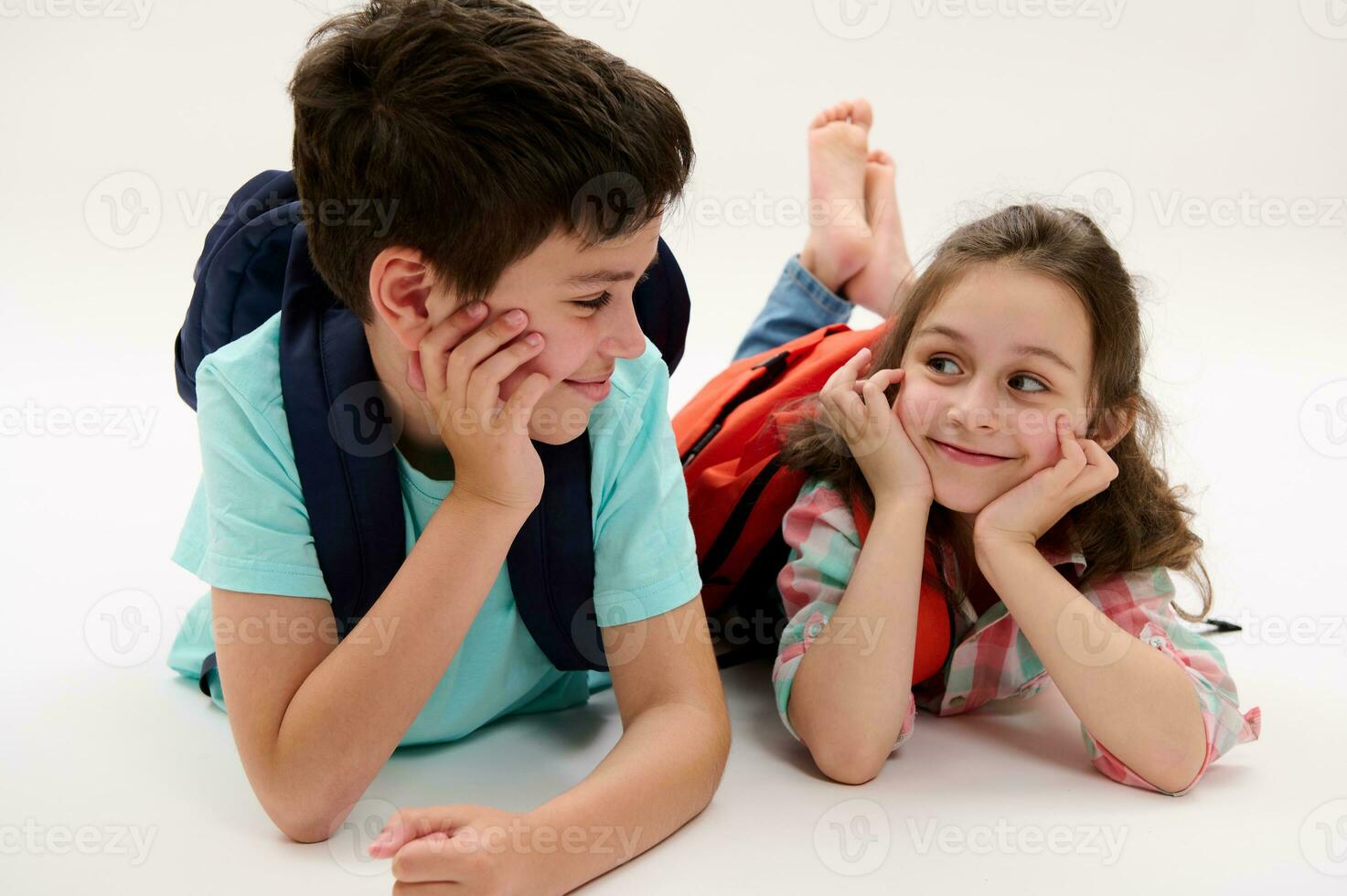 contento inteligente colegio niños, hermano y hermana con mochilas escolares, acostado en su barriga, aislado en blanco estudio antecedentes foto