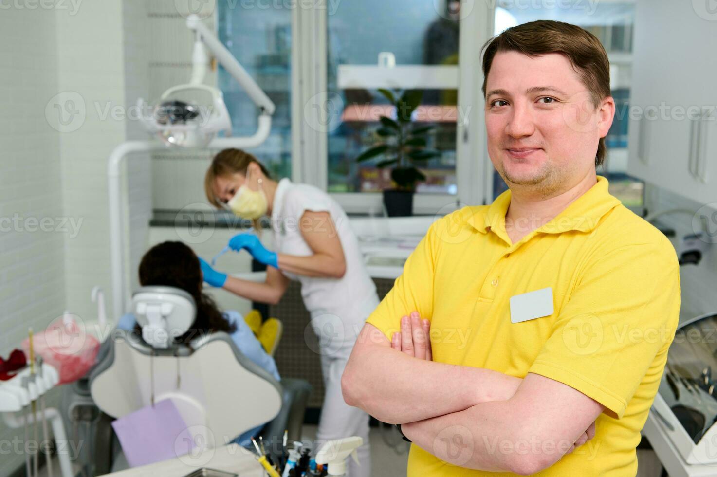 Confident happy male dentist in yellow uniform standing in dental office with arms folded, smiling looking at camera photo