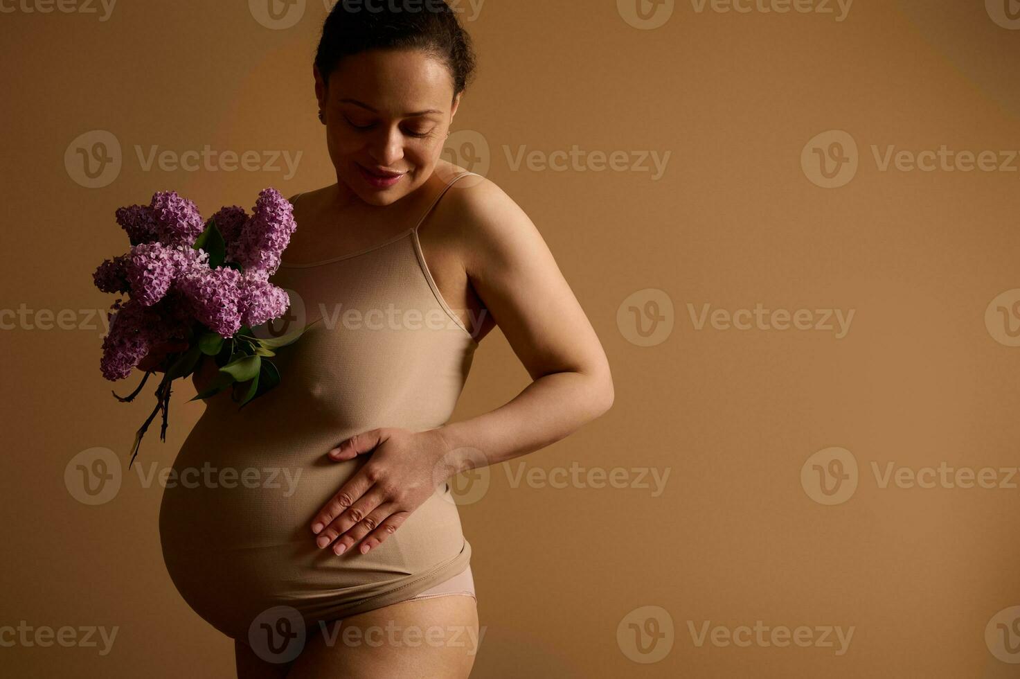 Attractive pregnant woman smiling, touching her belly, posing with a bunch of blooming lilacs, dressed in beige lingerie photo