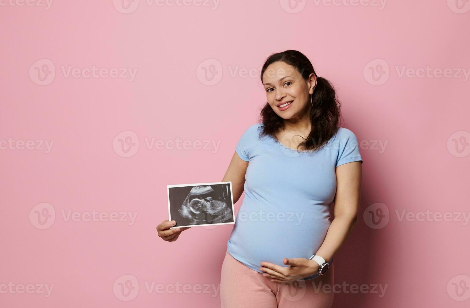 Gravid woman smiling at camera, holding baby ultrasound scan and caressing her pregnant belly, isolated pink background photo