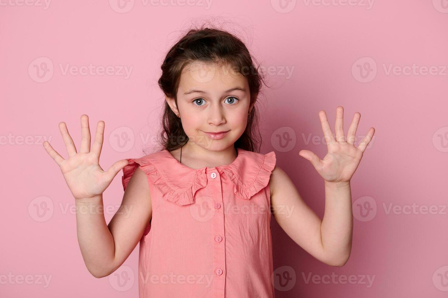 Studio portrait of coquettish sweet baby girl, lovely child showing hands palms to the camera, isolated pink background photo