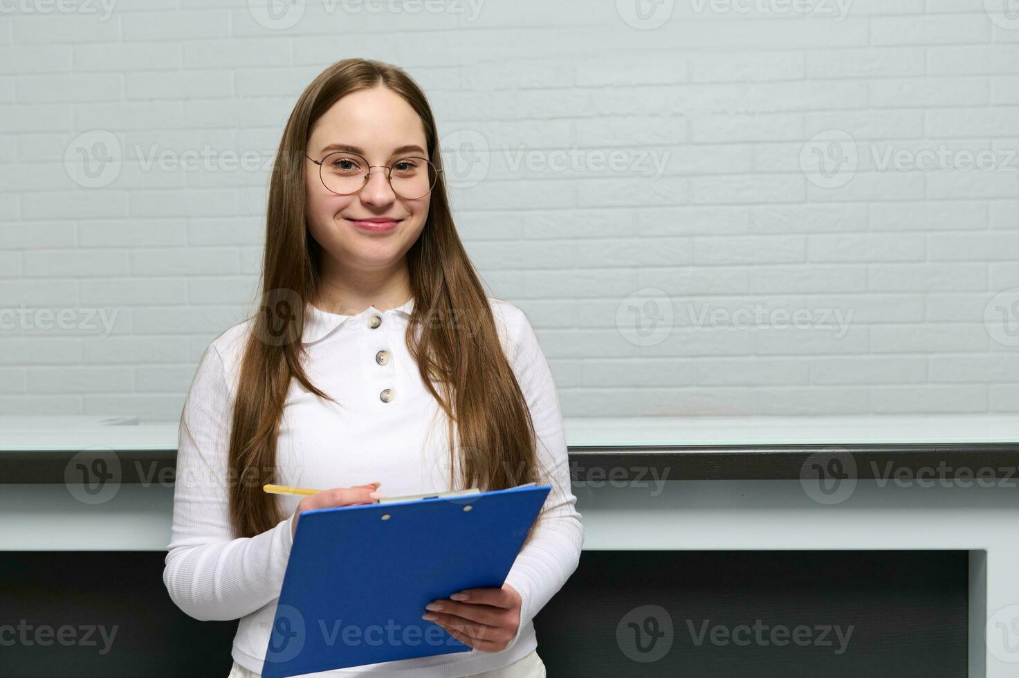 Confident portrait of a young woman, receptionist with clipboard near desk in modern clinic, smiling looking at camera photo