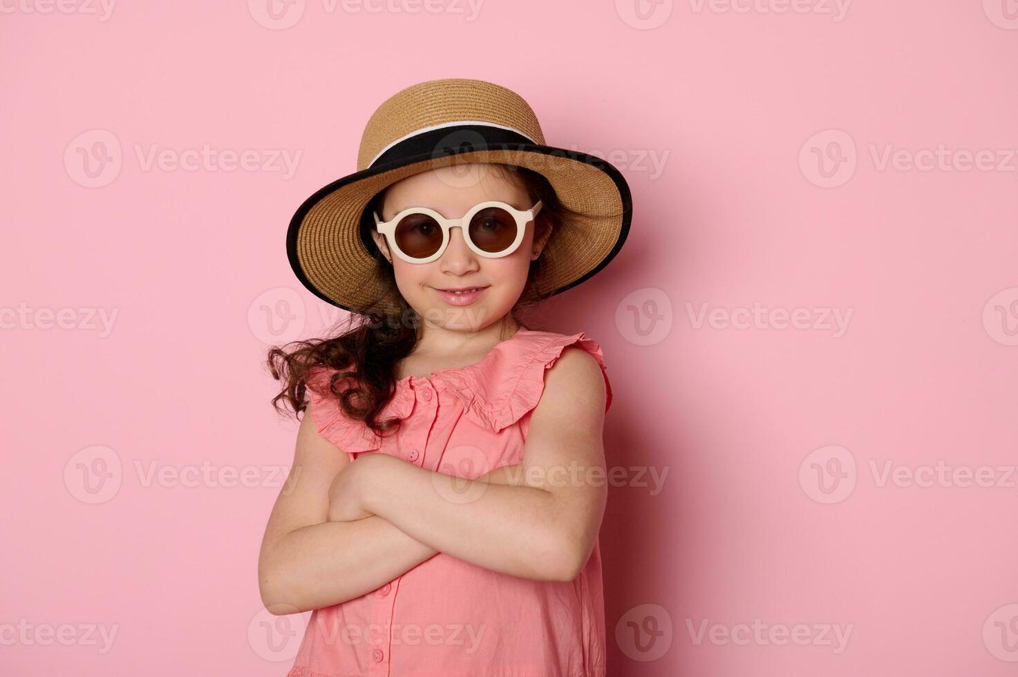 dañoso niño niña en Gafas de sol, rosado vestir y Paja sombrero, sonriente y posando con su brazos doblada en rosado antecedentes foto