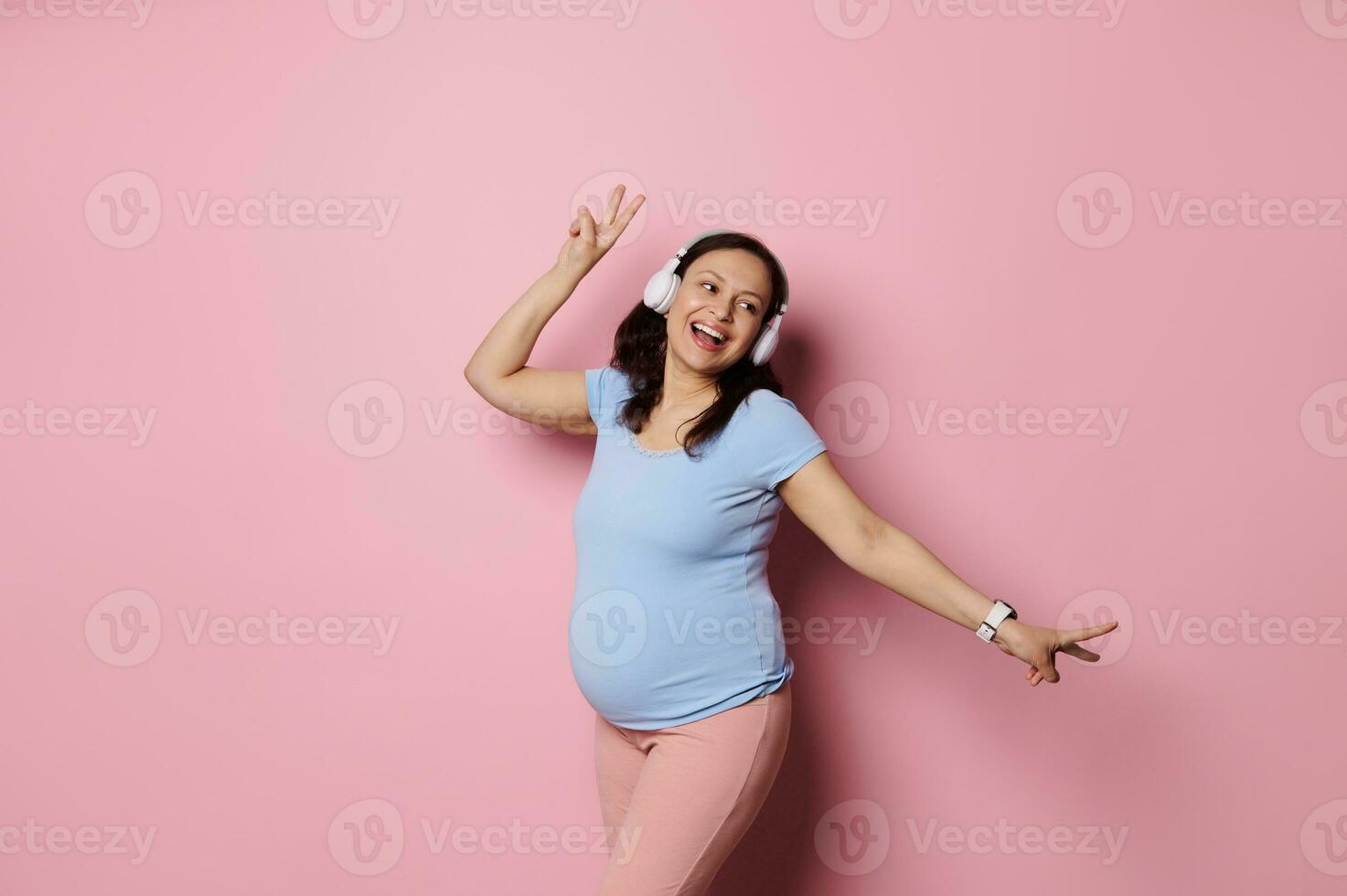 Joyful Latin American pregnant woman listening to music in wireless headphones and dancing over pink studio background photo