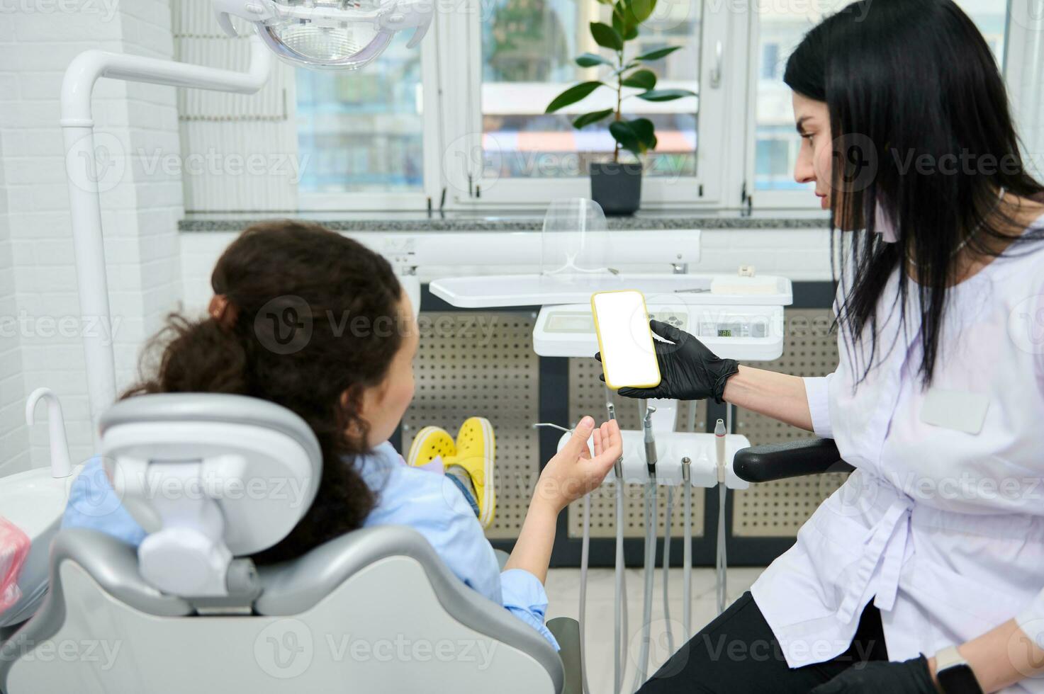 Rear view woman dentist holding smartphone with white blank screen and patient at dental appointment in dentistry clinic photo