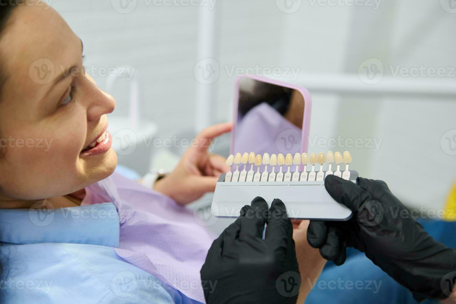 Smiling female patient receives dental care, teeth bleaching. Dentist checks tooth color using verneer Vita scale chart photo