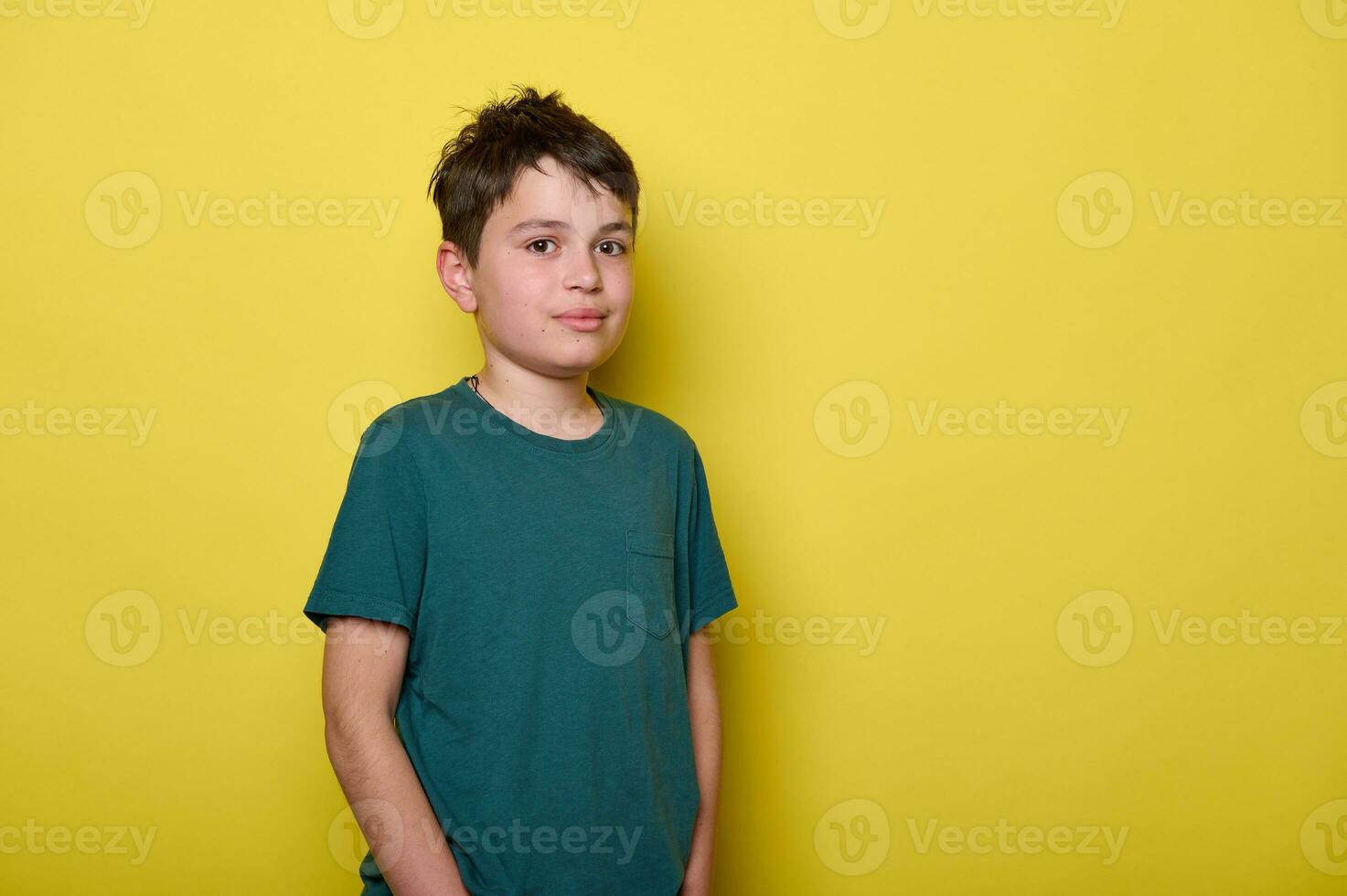 Handsome male teenager boy smiles looking at camera, standing on isolated yellow background with his hands in pockets photo