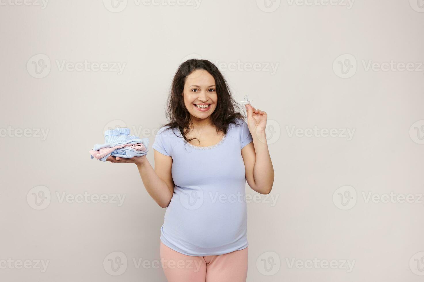 Happy pregnant woman, expectant mother holding clean laundered baby clothes, smiling looking at camera, isolated white photo