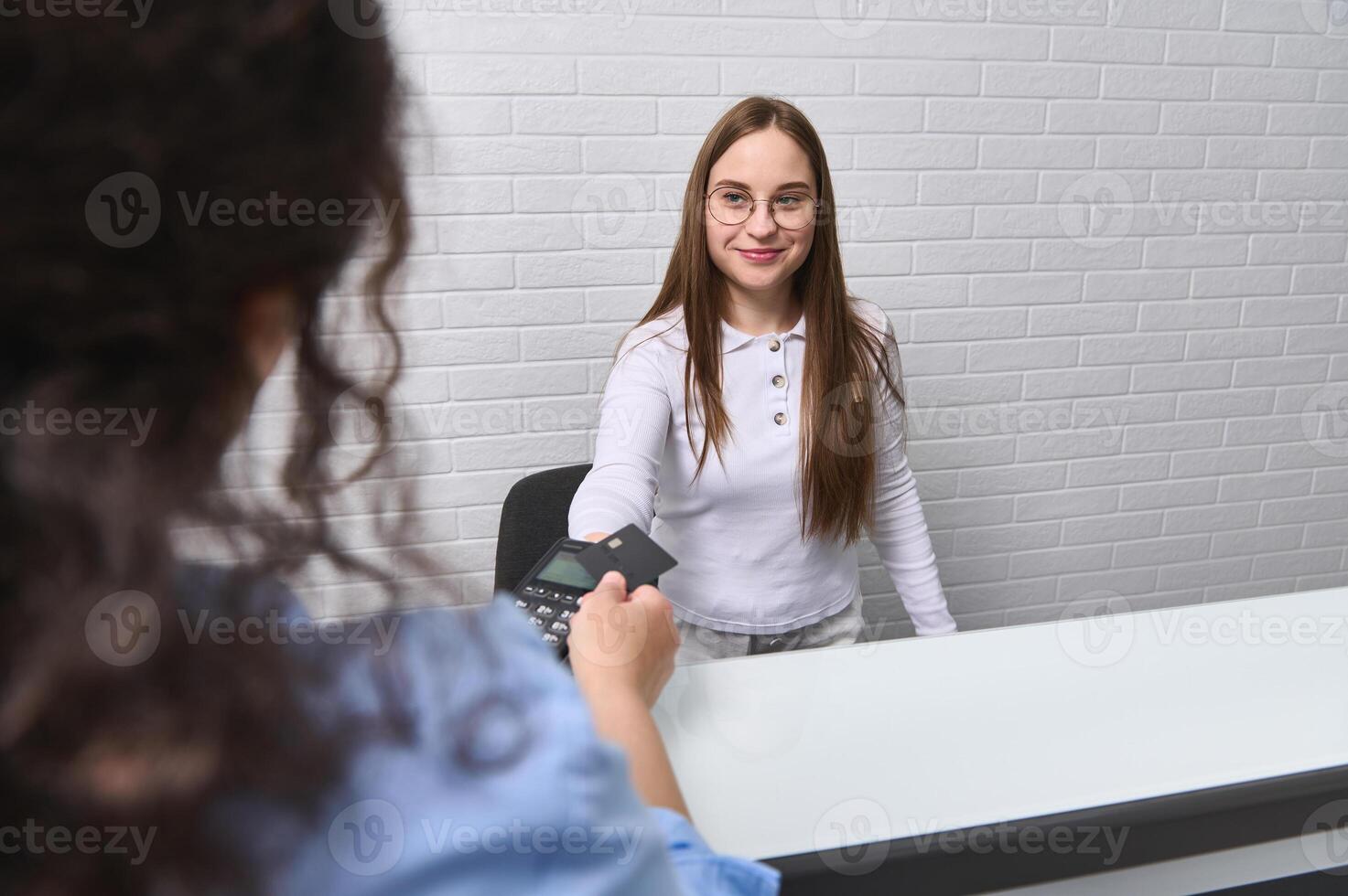 Smiling receptionist accepts cashless payment, holds out pos terminal to client for payment with credit card and NFC photo