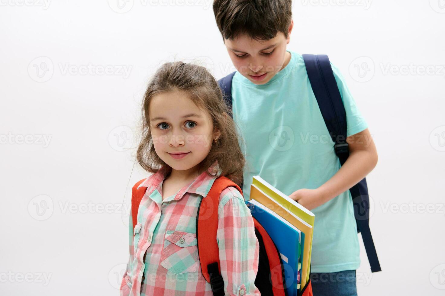 Happy little girl, first grader smiling looking at camera, and teen boy brother putting on workbooks inside her backpack photo