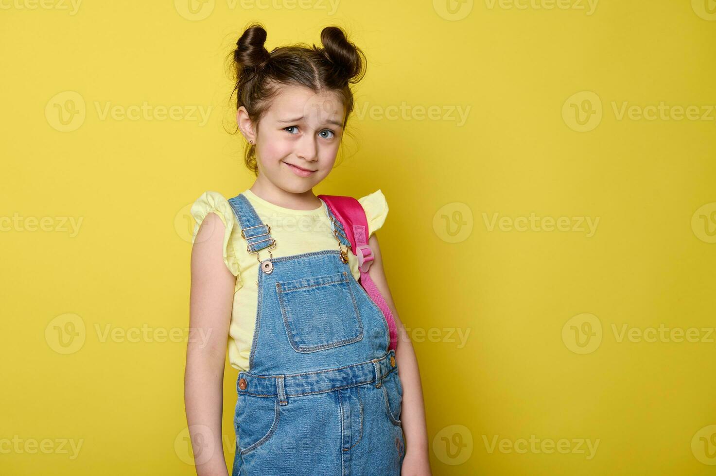 Emotional doubtful little kid girl, primary school student, pupil with pink backpack, yellow color studio background photo