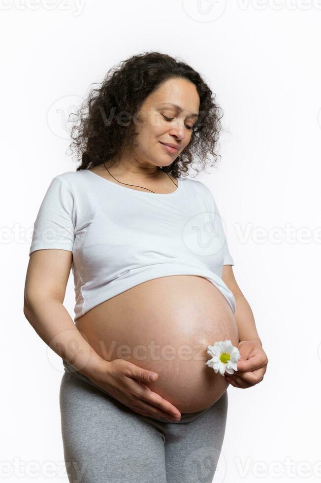 Lovely pregnant woman holding white daisy flower near her belly, gently stroking him over isolated studio background photo