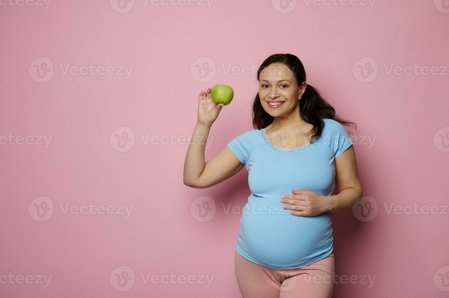 hermosa multiétnico embarazada mujer sonriente a cámara, conmovedor su barriga, participación un manzana, aislado rosado antecedentes foto