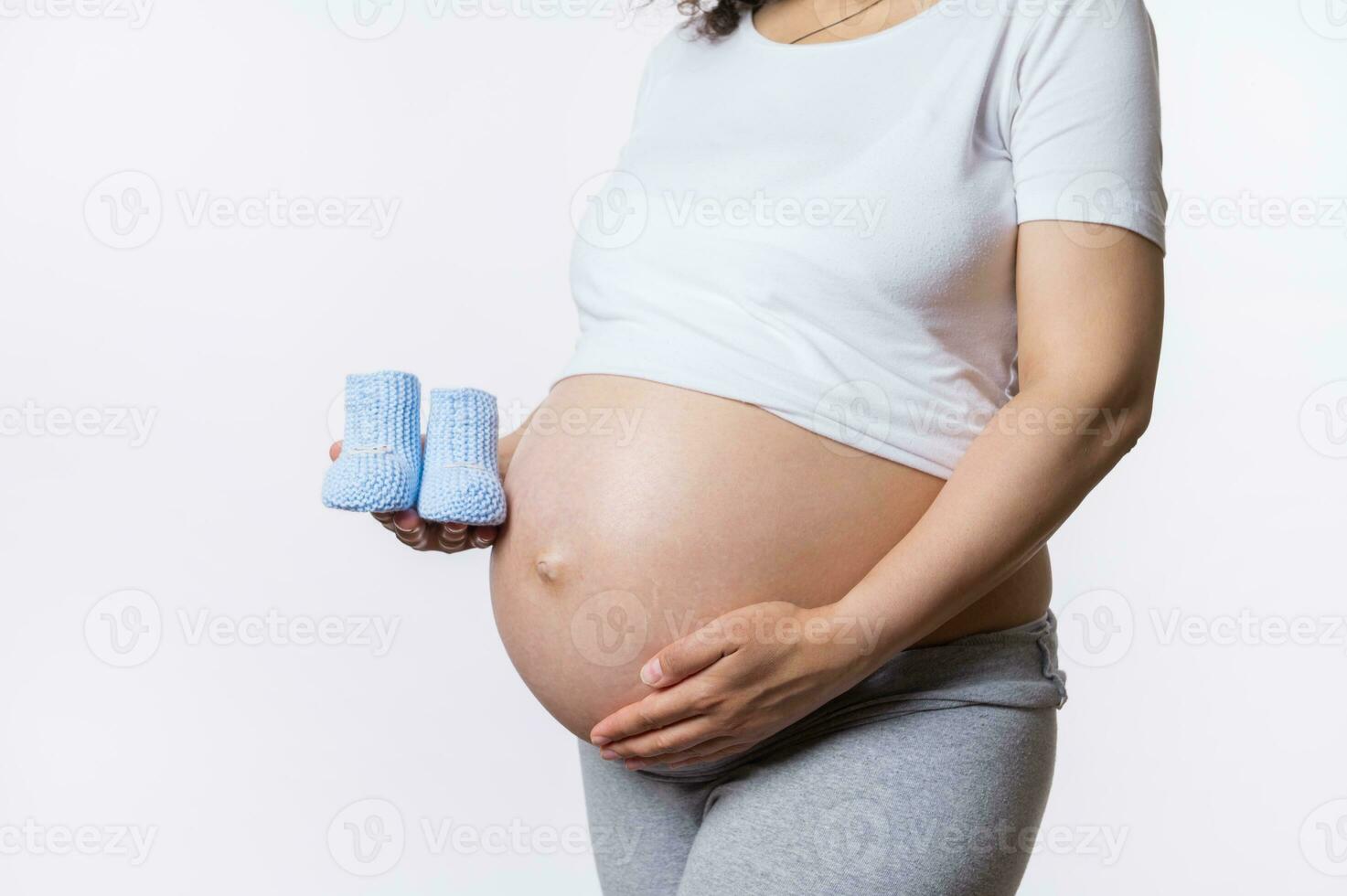 sección en medio de embarazada mujer conmovedor su grande barriga y participación azul de punto bebé botines, aislado en blanco antecedentes foto