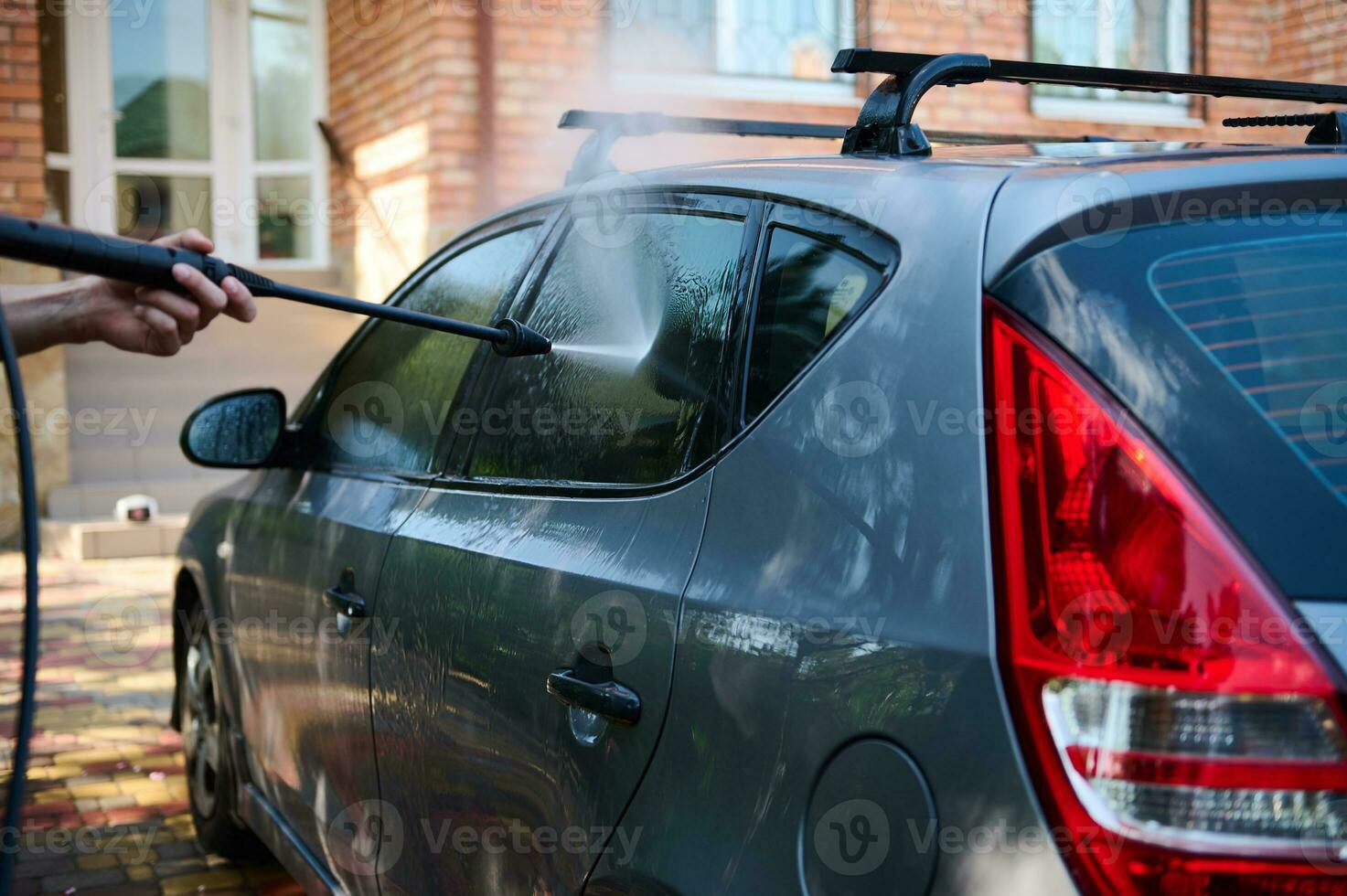 Close-up man washing car outdoors using a high pressure washer, spraying a pressurized water jet on the passenger window photo