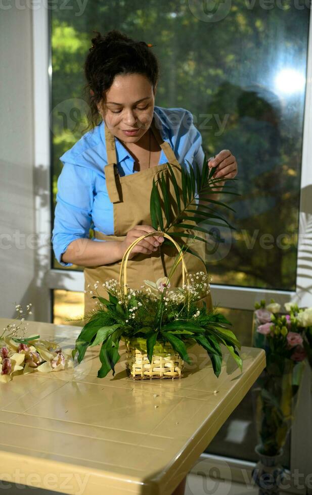 Female florist arranging a flower bouquet, inserting palm leaves into soaked foam in a wicker basket in floral shop photo