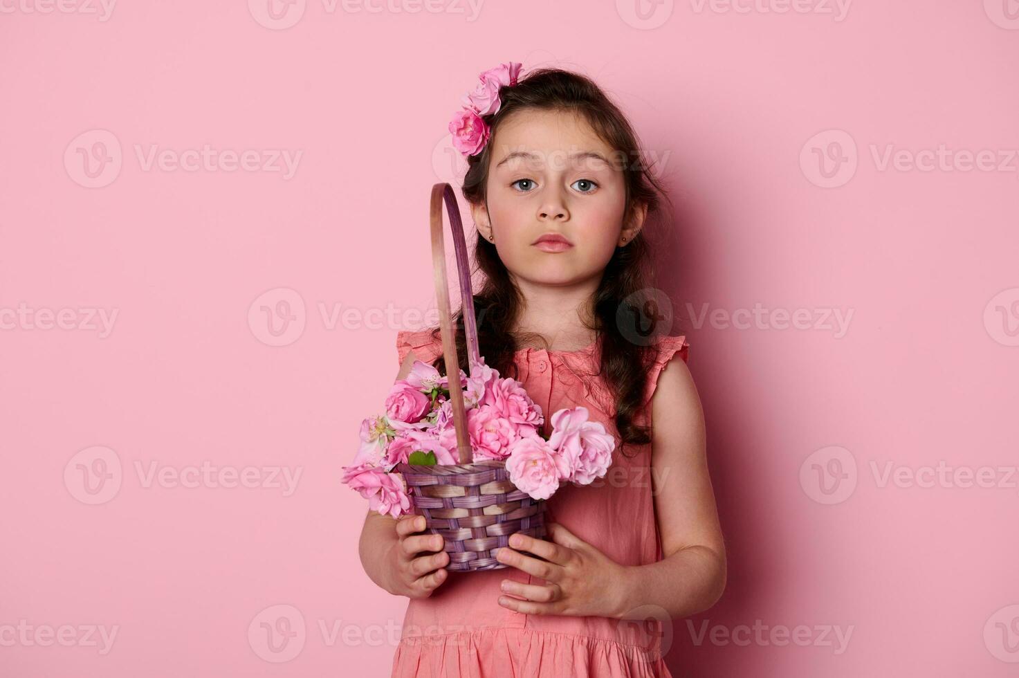 Noble little girl with curly hair and beautiful blue eyes, holding basket of pink roses, looking insightfully at camera photo