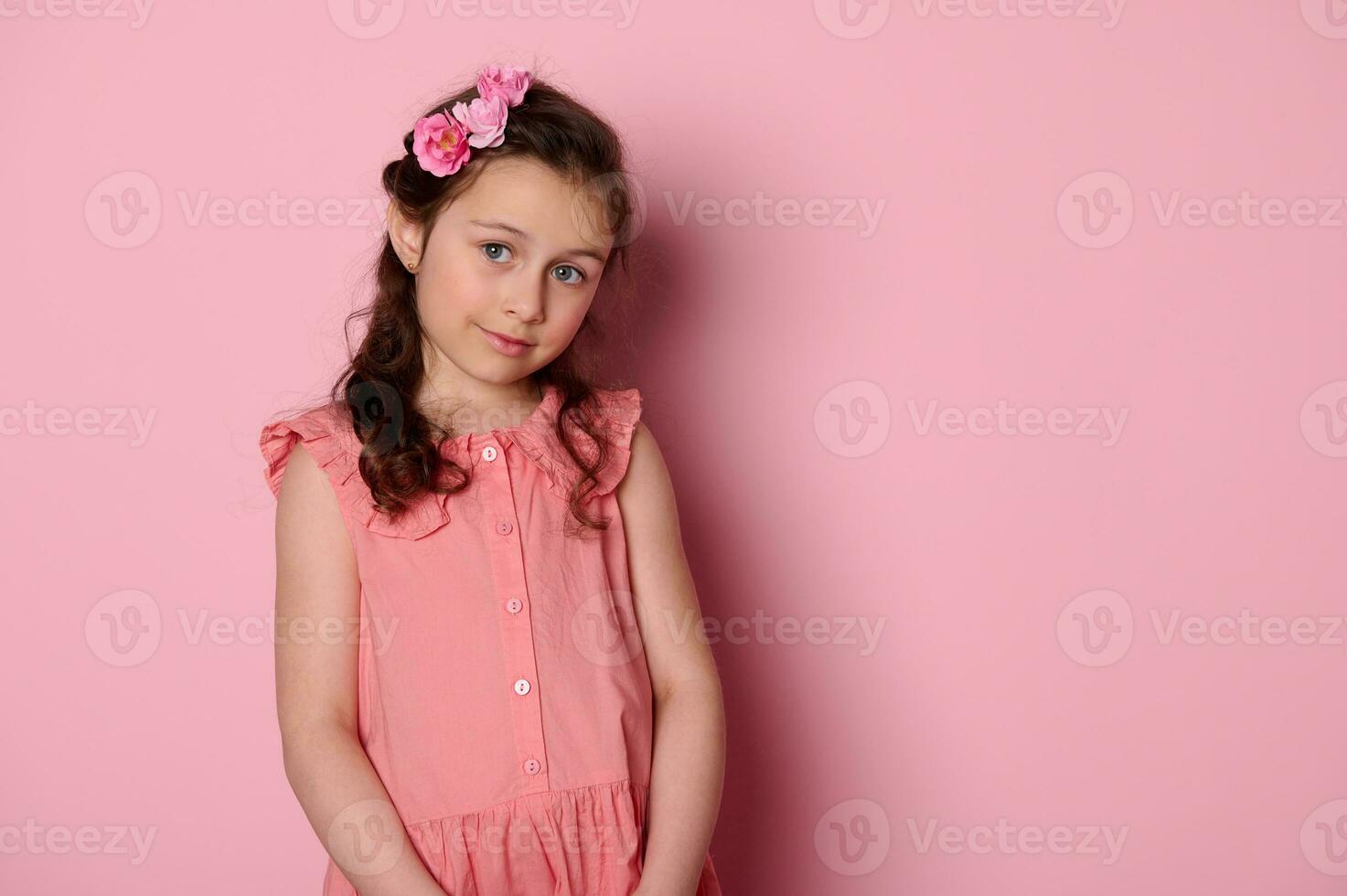 noble pequeño niña vistiendo rosado vestir y peinado con rosas, mira a cámara con perspicaz mirada, aislado antecedentes foto