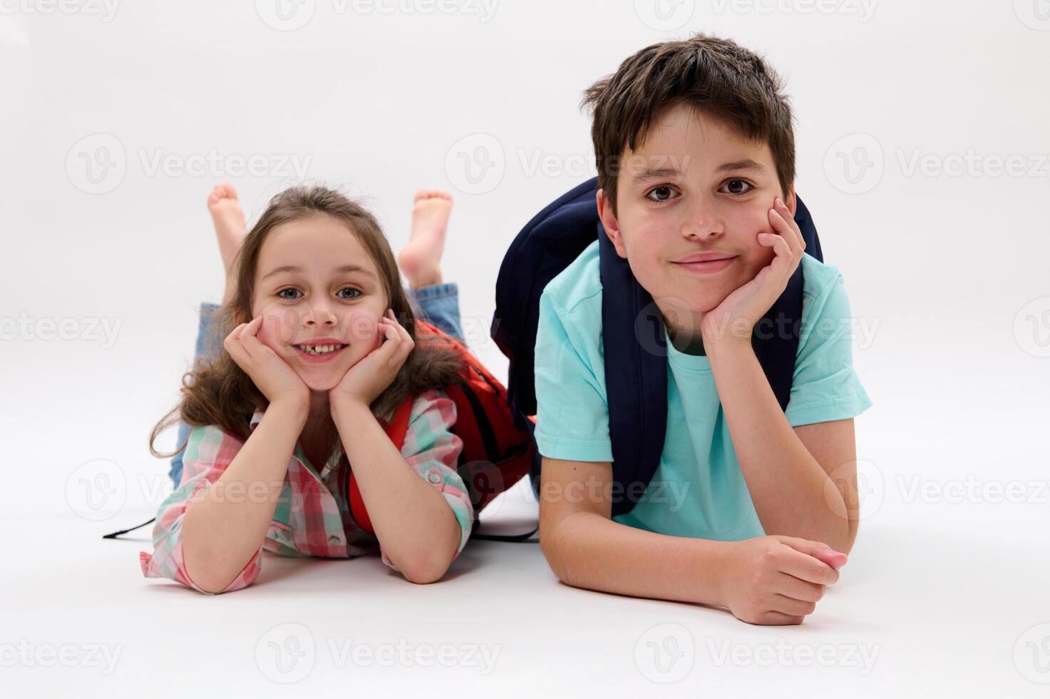 Smart school children, first grade kid girl and teenage schoolboy, lying on belly, smiling at camera, isolated on white photo