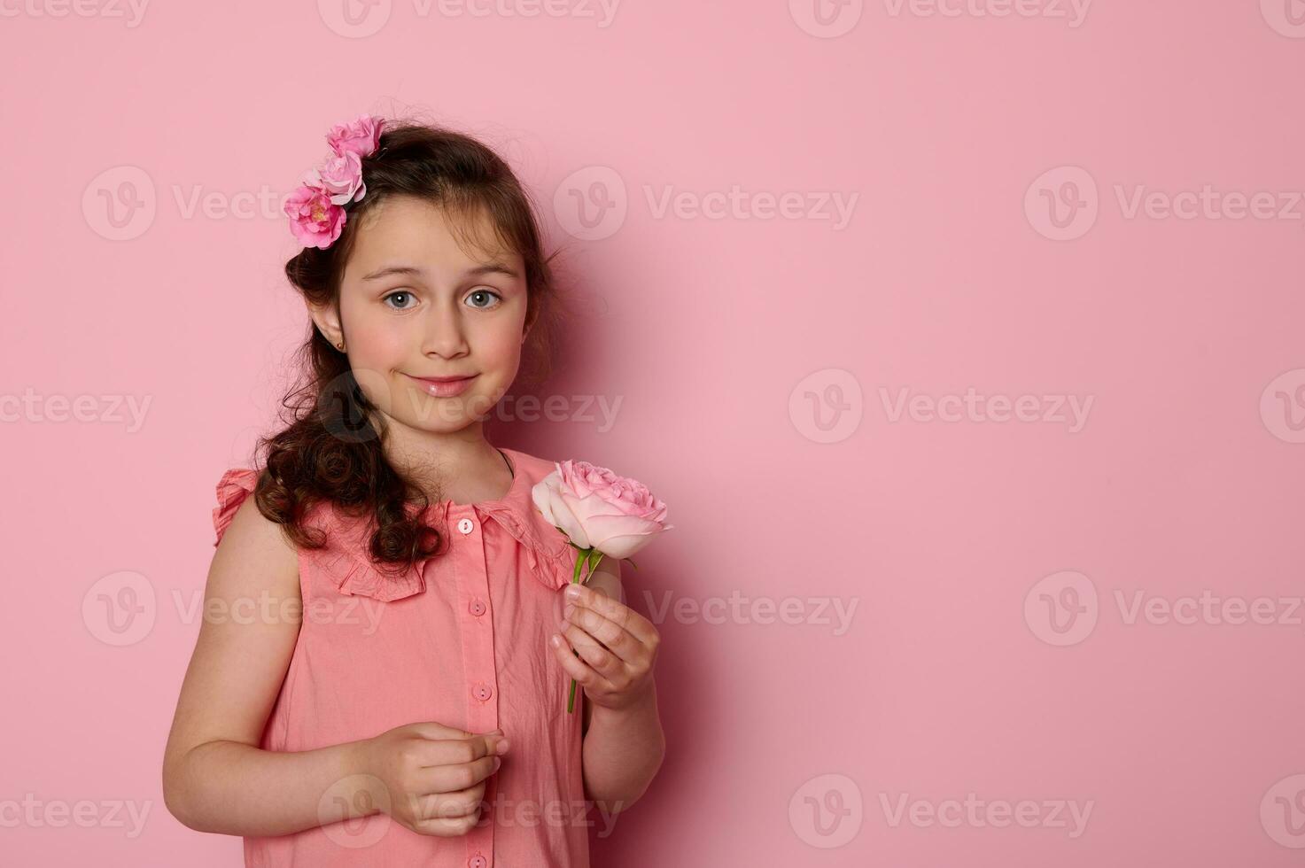 Caucasian lovely child girl in stylish pink dress, holding a beautiful rose flower, smiling on isolated pink background photo