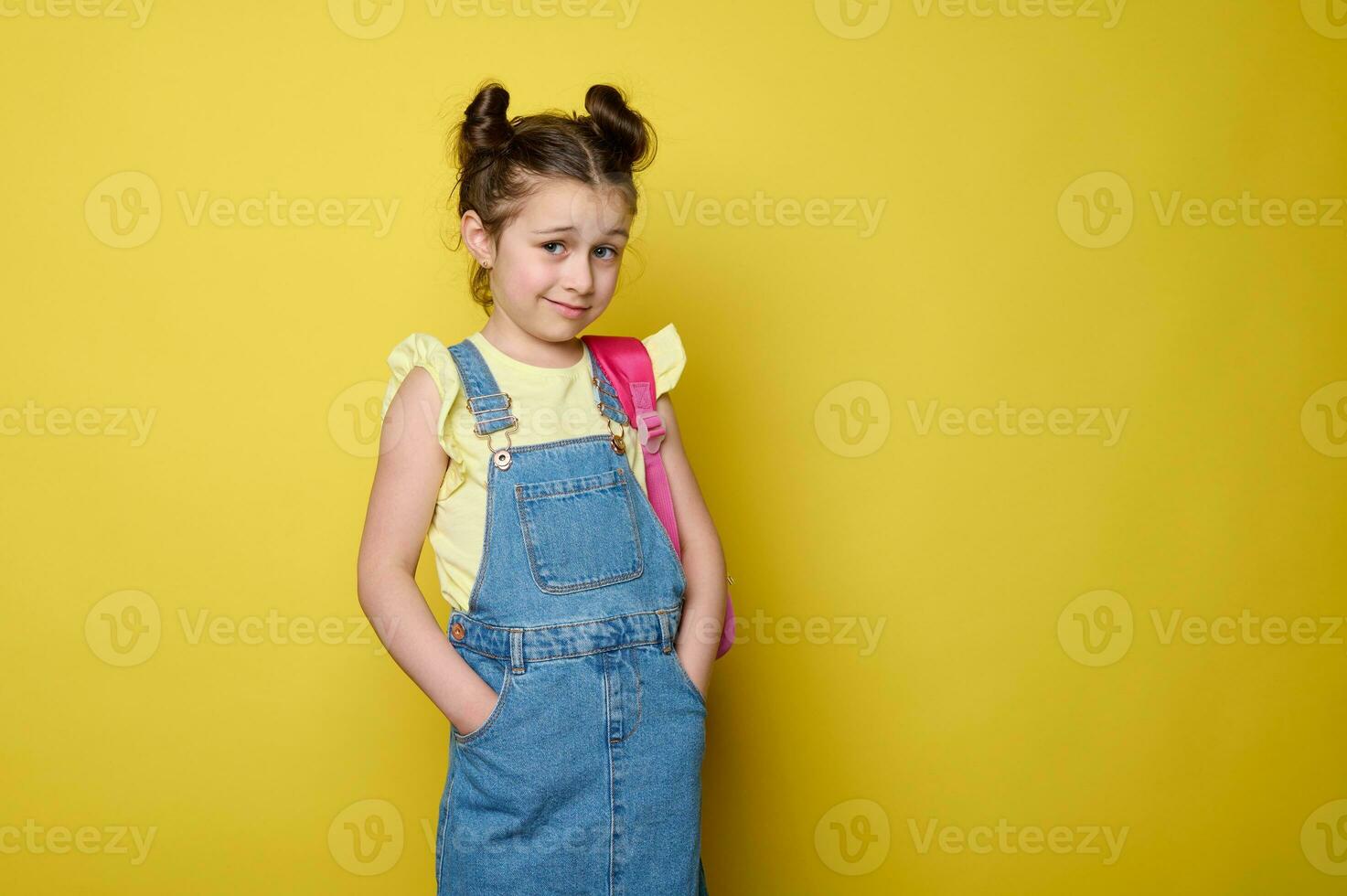 adorable colegio niña en azul mezclilla mono, que lleva un rosado mochila, posando con manos en bolsillos en amarillo antecedentes foto