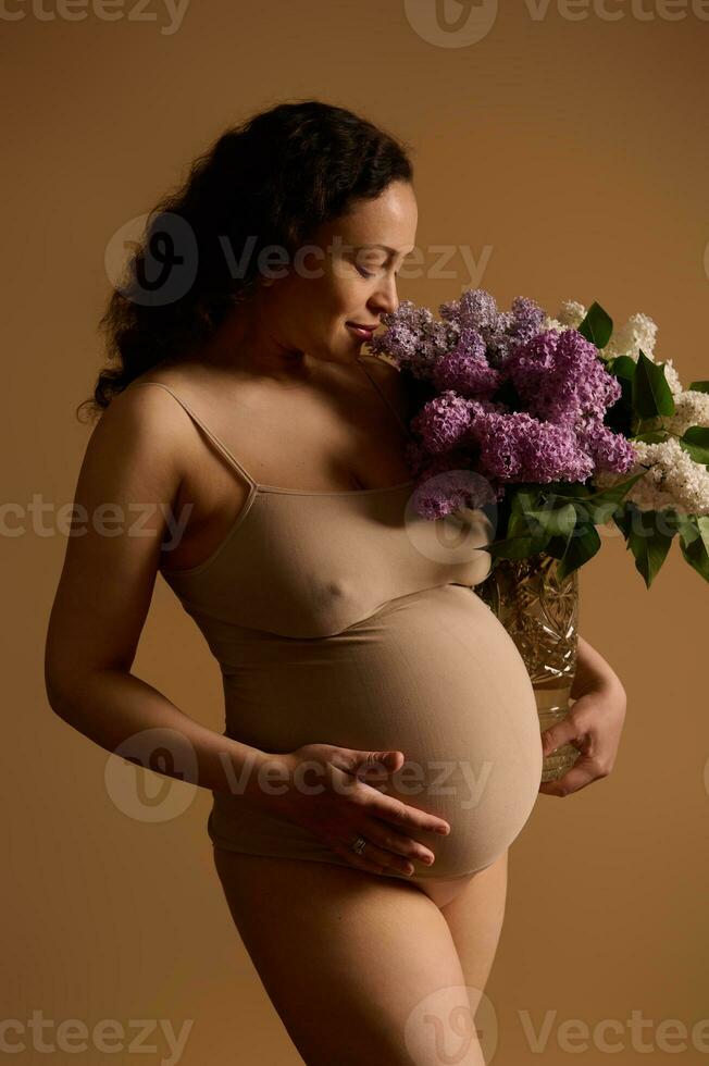 hermosa embarazada mujer en beige ropa interior, poses con manojo de lilas, aislado en beige antecedentes. el embarazo 30 semana foto